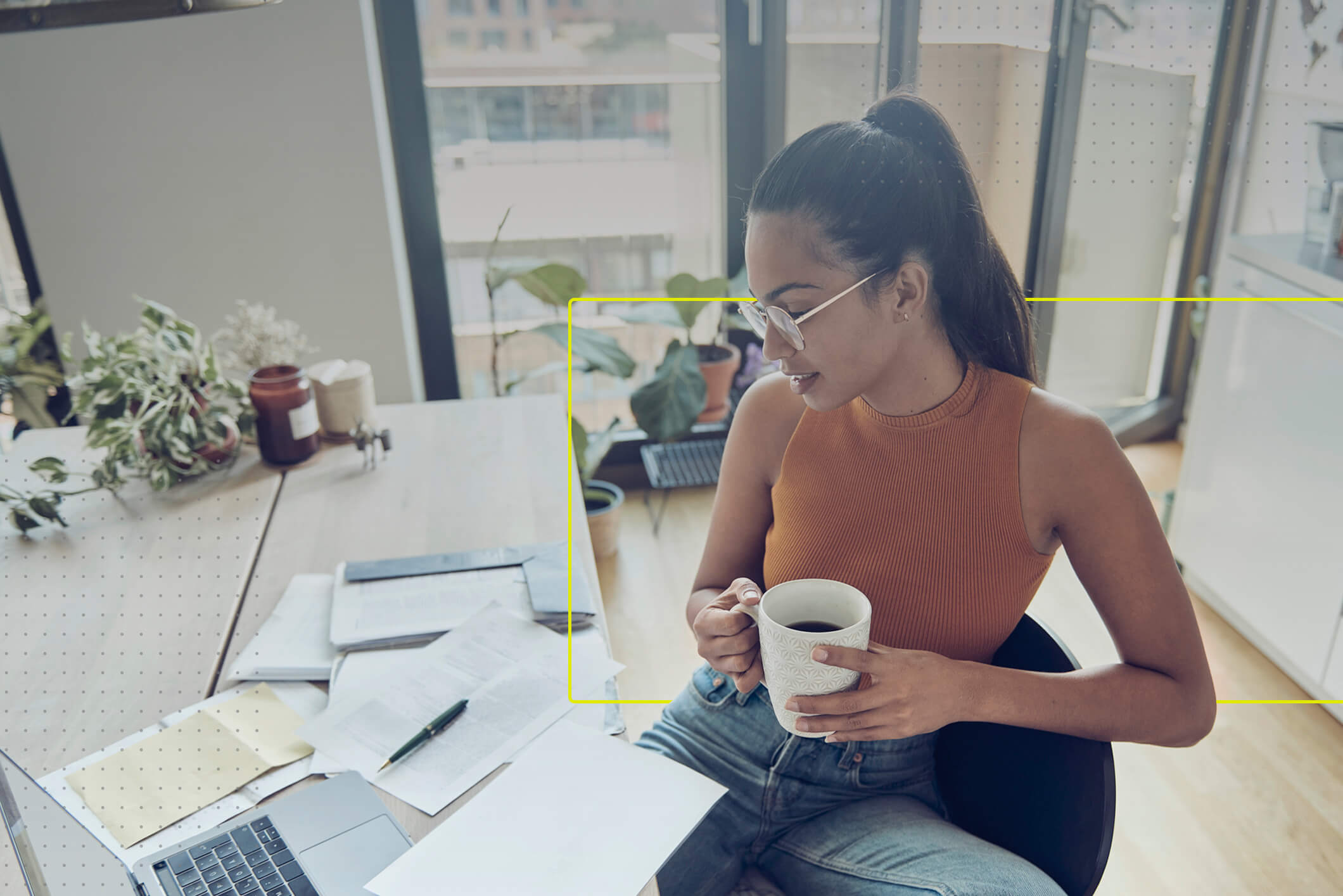 Una mujer de mediana edad sentada en la sala de un departamento revisa su computadora mientras bebe una taza de café.