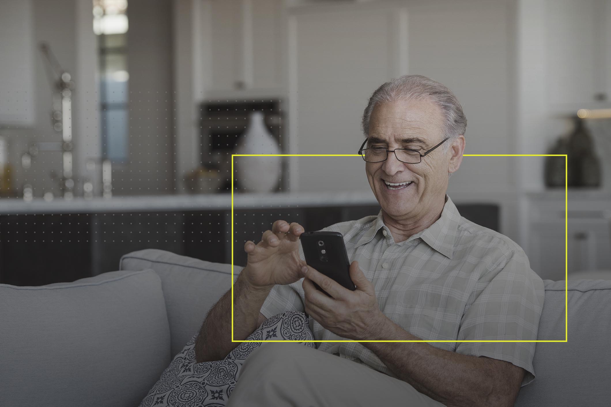 Una persona, hombre adulto mayor, revisando su teléfono inteligente sentado en un sillón de la sala de su casa.