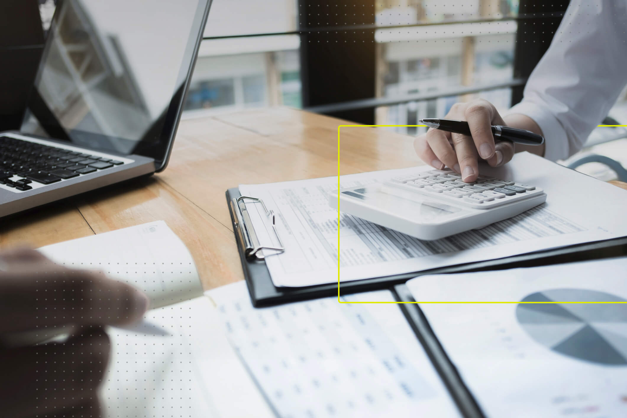 Un escritorio en primer plano, con varios documentos financieros y dos personas de frente sentadas. Una persona tiene una laptop, la otra una calculadora.